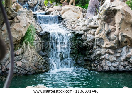 Very beautiful artificial waterfalls with living water and growing moss. Water flows from above, splashes and drops around. Open spaces and parks for walking. Summer period, cool pond.
