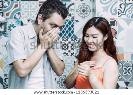 Young couple standing in bathroom and looking at pregnancy test. Woman is smiling. Man is shocked.