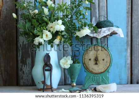 Floral composition with old rusty vintage kitchen scale, lace, sisal twine on bowl, bunch of keys, roses in clay pot, scissors in vase, rustic secateurs on weathered wooden background, vintage style