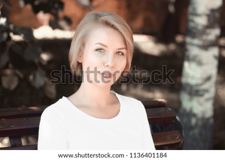 Portrait of young blonde girl with short hair looking at camera and sitting on bench in park in white shirt.