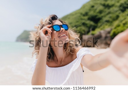 Cheerful girl with tanned skin making selfie at tropical island. Outdoor photo of ecstatic young woman in trendy sunglasses taking picture of herself at sandy beach.