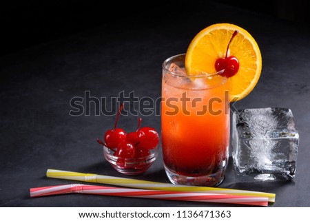 Tequila sunrise cocktail. Maraschino cherry, straws and ice cube on a dark stone background with space for your text.