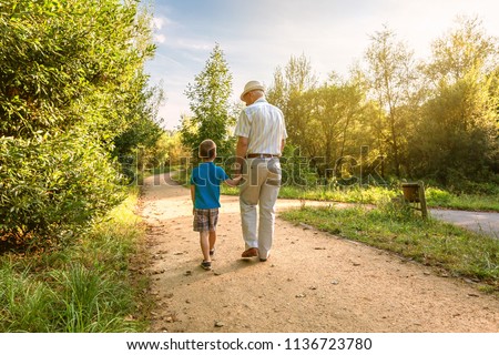 Back view of grandfather with hat and grandchild walking on a nature path Royalty-Free Stock Photo #1136723780