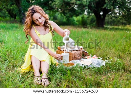 a girl in a yellow dress pours milk from a white jug to the white cup. picnic basket in the background. 