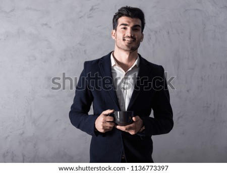 A happy handsome young businessman in a black suit holding a black mug in front of a grey wall in a studio.