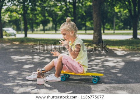 side view of little child licking fingers from dessert and sitting on skateboard at urban street