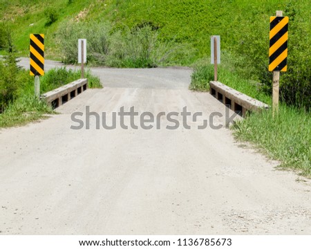 rural gravel road leading over narrow wooden bridge with yellow and black caution signs near a lush green hillside in Montana
