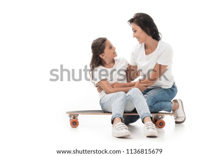 mother sitting near smiling daughter on skateboard isolated on white