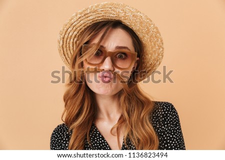 Portrait of amusing funny woman 20s wearing straw hat and sunglasses fooling around and putting hair like mustache isolated over beige background
