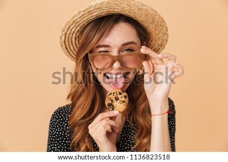 Image closeup of lovely excited woman 20s wearing straw hat and sunglasses eating biscuits isolated over beige background