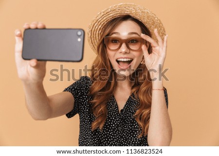 Portrait of cheerful pretty woman wearing straw hat and sunglasses laughing and taking selfie photo on smartphone isolated over beige background