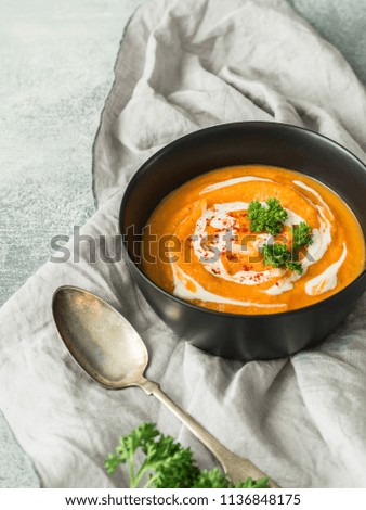 Pumpkin cream soup with cream, dry paprika and fresh parsley in a black bowl on a kitchen towel on a gray background