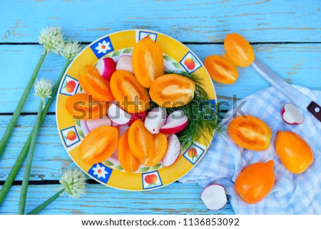 flat lay on a blue painted old wooden table with fresh dill and onion greens, cucumbers and orange tomatoes with radishes, sliced in a plate, cotton caged napkin and knife.