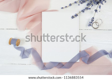 White blank card and ribbon with two wedding rings on of pink fabric with lavender flowers on a white background. Mockup with envelope and blank card with space for your text. Flat lay. Top view