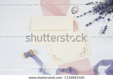 White blank card, envelope and ribbon with two wedding rings on a background of pink fabric with lavender flowers on a white background. Mockup with envelope and blank card. Flat lay. Top view