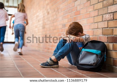 Little boy sitting alone on floor after suffering an act of bullying while children run in the background. Sad young schoolboy sitting on corridor with hands on knees and head between his legs. Royalty-Free Stock Photo #1136889179