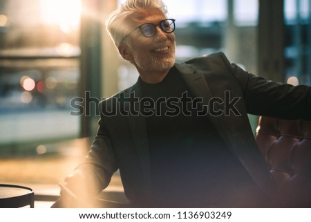 Smiling mature businessman sitting in office lobby with bright light in background. Happy senior man in business clothes sitting in office lounge.