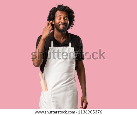 Handsome african american baker crossing his fingers, wishes to be lucky for future projects, excited but worried, nervous expression closing eyes