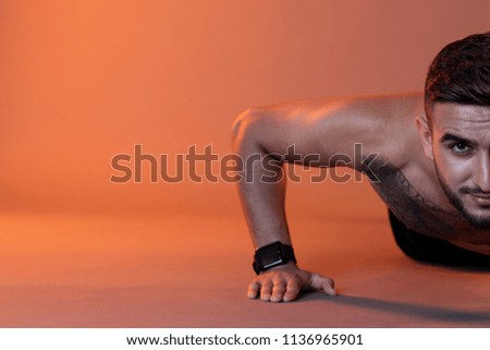 Crop tattooed shirtless man looking at camera while doing push-ups on floor against red illuminated background