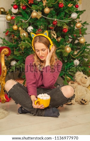 cute teenager girl sitting on the floor near big Christmas tree and listening the music and drink her cacao with marshmallow alone