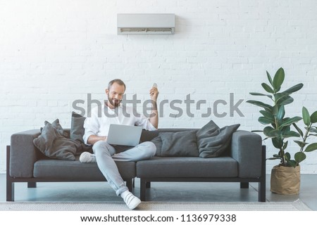 man turning on air conditioner with remote control while using laptop on sofa Royalty-Free Stock Photo #1136979338