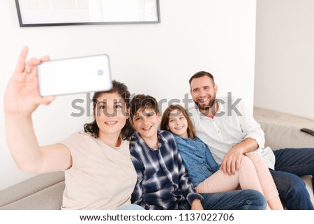 Picture of joyous content family with two children sitting on sofa together at home and taking selfie photo on smartphone
