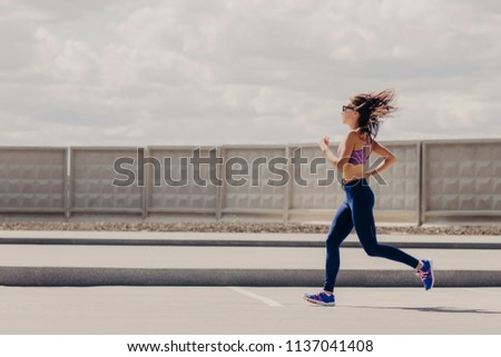 Freeze action of beautiful slim woman has morning jogging exercise, wears top and leggings, breathes fresh air while runs, enjoys sport, has healthy lifestyle. Female runner has workout outdoor