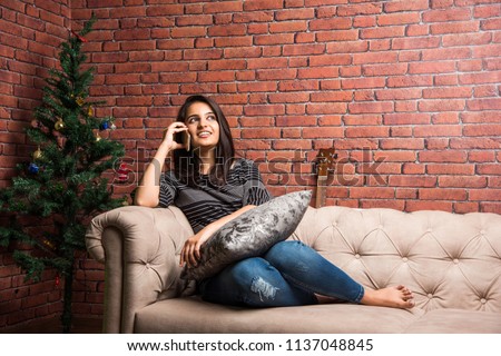 Pretty Indian / Asian girl using smartphone while seating on sofa or couch at home
