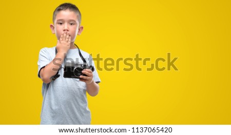 Dark haired little child holding vintage camera cover mouth with hand shocked with shame for mistake, expression of fear, scared in silence, secret concept