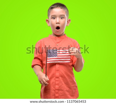 Dark haired little child holding United States flag scared in shock with a surprise face, afraid and excited with fear expression