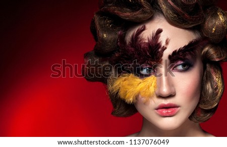 Fashion unusual girl with big curls. Bright creative makeup, red and yellow feathers on the face. Isolated on black background