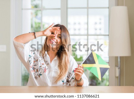 Young woman at home holding flag of Jamaica with happy face smiling doing ok sign with hand on eye looking through fingers