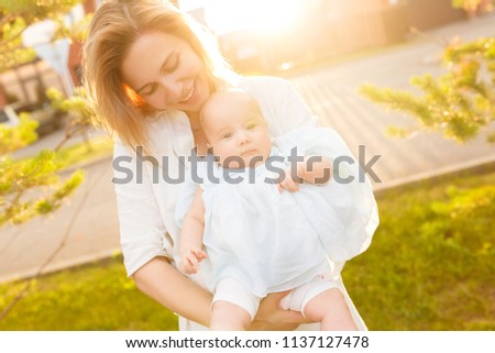 Close up cropped shot of half-year little baby girl in arms of sweet mommy. Cute child looking at camera. Soft sun light picture. Happy family spend time together in summer, white clothes