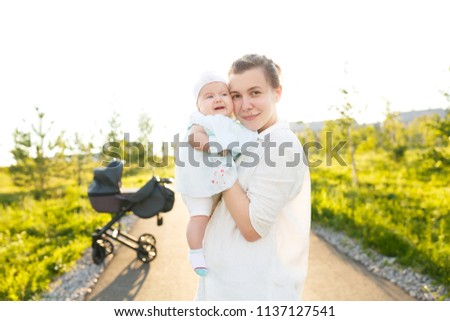 Loving family. Nice picture of charming young mommy in white casual clothes hold little baby girl in arms, gently embrace child over green meadow, nature background at sunset evening.
