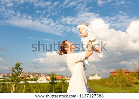 Happy family shot. Beautiful picture of ypung attractive mommy holds toddler upstairs, has hice mood, rejoices at motherhood on beauty sky blue background with clouds and green city park outdoor
