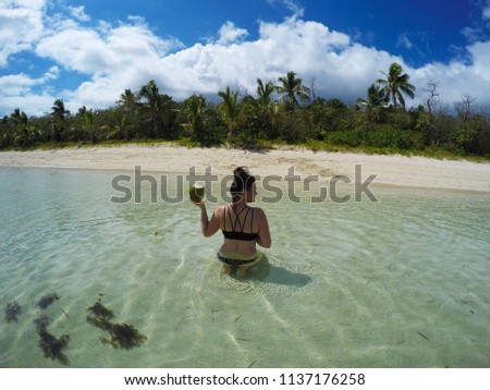 Travel vacation concept background. Young tourist woman sitting on exotic beach in the clear water holding coconut cocktail in her hand. Fiji Islands, Pacific. Bright colours