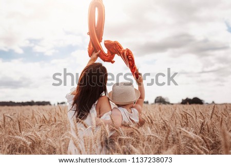 Mother with a toddler from behind in corn field holding red balloon.