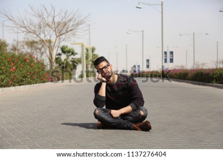 Sad man sitting crossed legs on the street puts hand on cheek, on the road of the cars in the mall.
