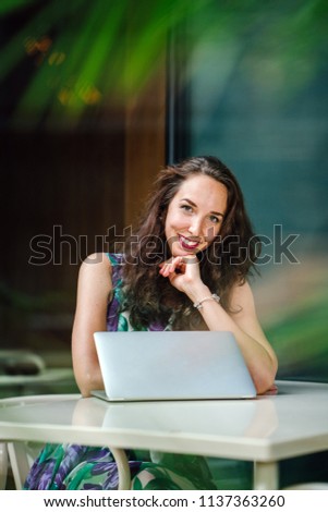 Portrait of a young Caucasian woman executive working on her laptop computer at a table outdoors during the day. She is sitting at a cafe, coworking space or office and is smiling happily as she works