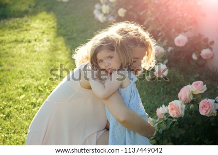 Woman with girl child at blossoming rose flowers. Mom cuddling daughter on green grass on sunny summer day. Family love and care. Mothers day concept. Childhood and parenting.