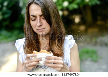 Beautiful woman with dark hair eating fast food hamburger outdoor. Feelings bad with stomach pain