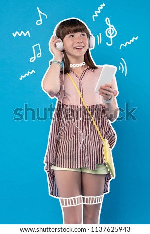 Amazing music. Cheerful positive teenager standing with a modern device and listening to music in her big headphones
