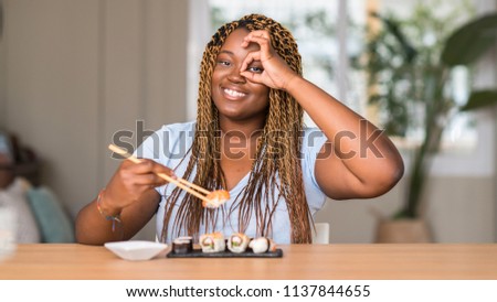 African american woman eating sushi with happy face smiling doing ok sign with hand on eye looking through fingers