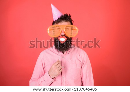 Guy in party hat celebrates, posing with photo props. Hipster in giant sunglasses celebrating. Man with beard on cheerful face holds smiling mouth on stick, red background. Photo booth fun concept.