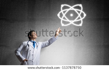 Successful and young doctor in white uniform interracting with molecular structure while standing against gray concrete wall on background.