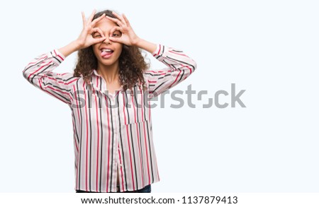 Beautiful young hispanic woman doing ok gesture like binoculars sticking tongue out, eyes looking through fingers. Crazy expression.