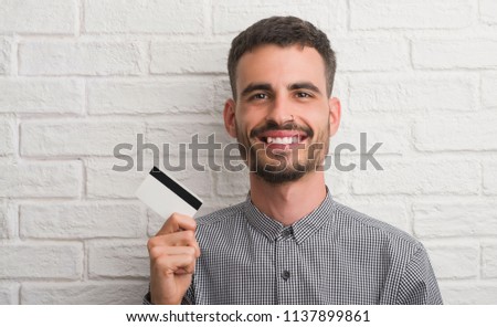 Young adult man over brick wall holding credit card with a happy face standing and smiling with a confident smile showing teeth