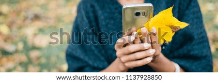 Detail of woman using her phone and holding autumn yellow leaves. Fall season mobile communication banner.