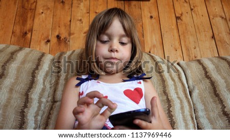 A child is sitting on the couch and playing a smartphone. A girl of European appearance, 4-5-6 years old with blond hair playing or studying a smartphone. Concept of play and learning.