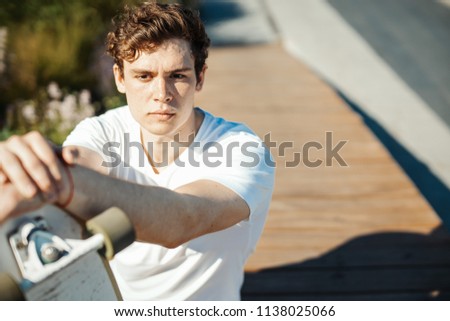 Young attractive man sitting on the bench in the park and holding longboard.